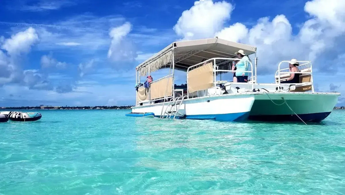 A white and blue catamaran with a tan canopy and fringe trim floats on crystal-clear turquoise water under a bright, cloudy sky