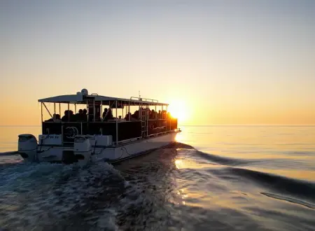 A boat filled with passengers travels across calm water toward a bright, glowing sun on the horizon