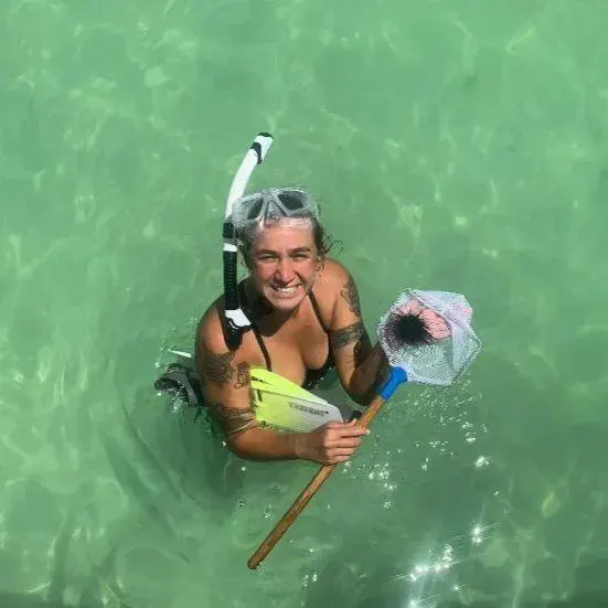 A smiling woman wearing snorkeling gear standing in clear water, holding up a small net with a sea urchin inside