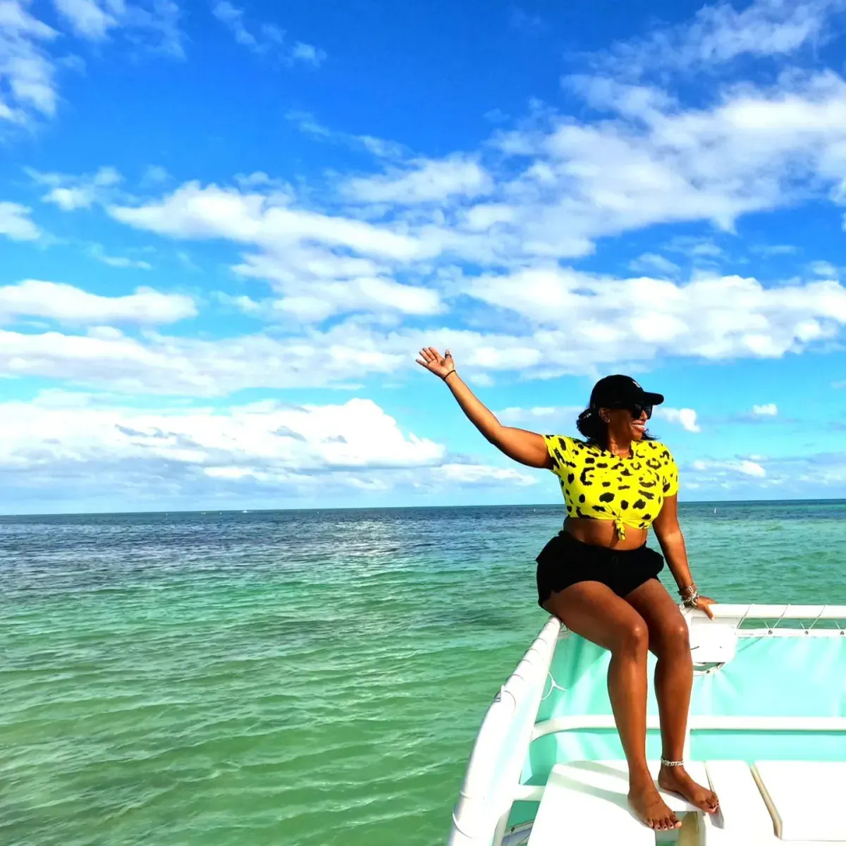 A woman in a yellow leopard-print top and black shorts sitting on the edge of a boat and waving toward the ocean