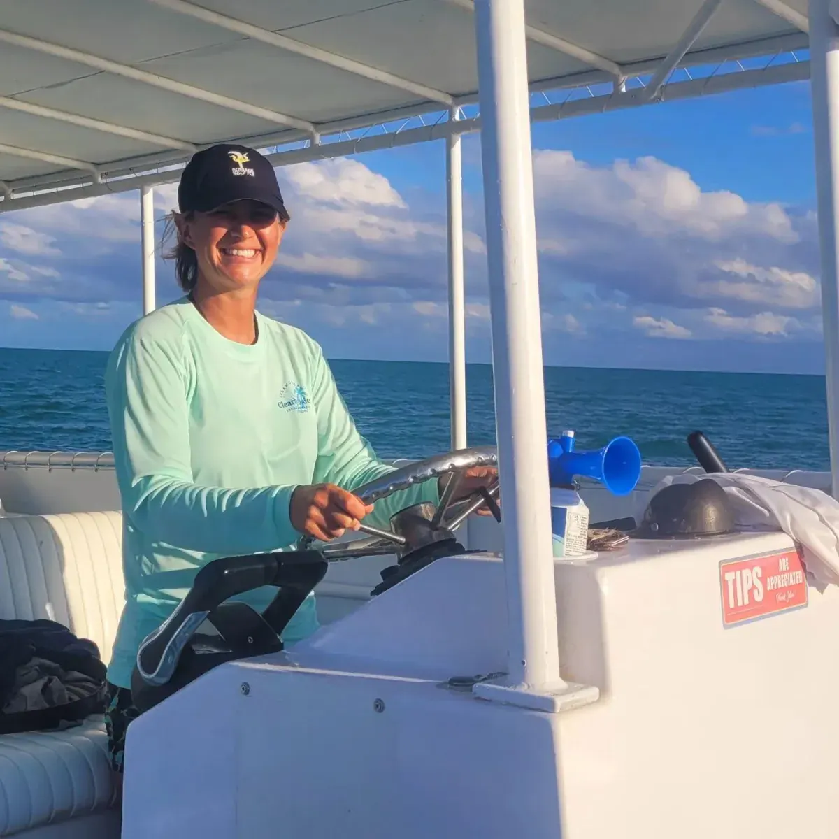 A smiling woman in a teal long-sleeve shirt and baseball cap steers a boat on a calm blue ocean under a cloudy sky