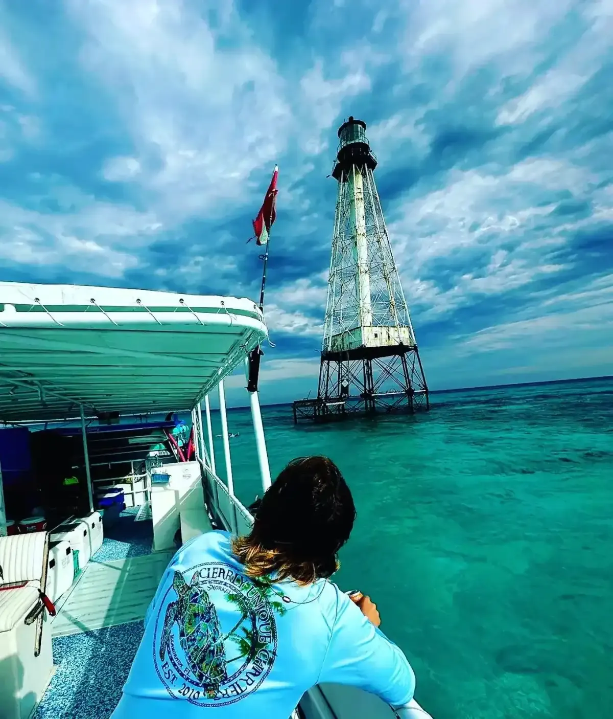 A person in a light blue shirt looks out from a boat toward a tall, skeletal lighthouse standing in vibrant turquoise water under a dramatic, cloudy sky