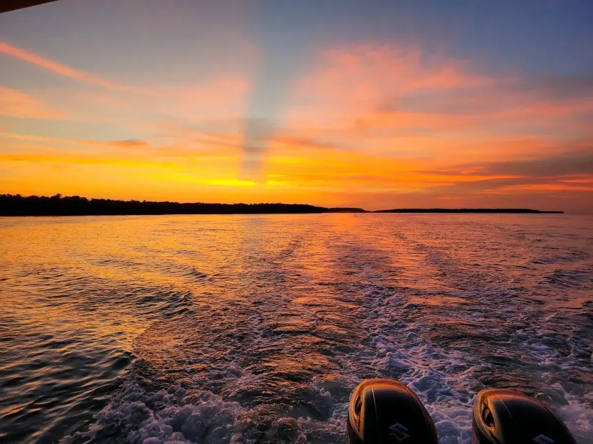 A view from the back of a moving boat showing a white wake trailing toward a vibrant orange and yellow sunset over a dark shoreline