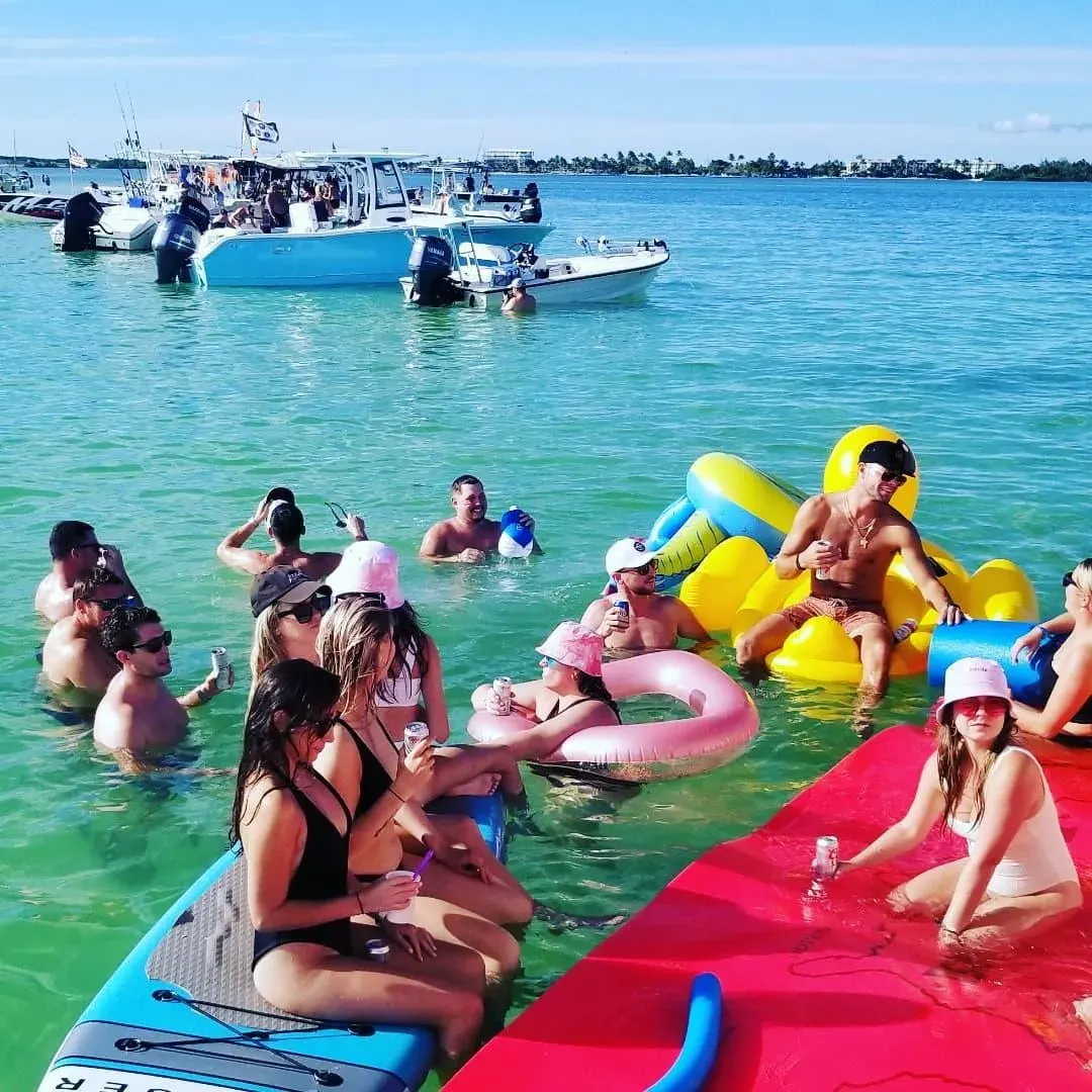 Group relaxing in shallow turquoise water with drinks, paddleboards and colorful floats