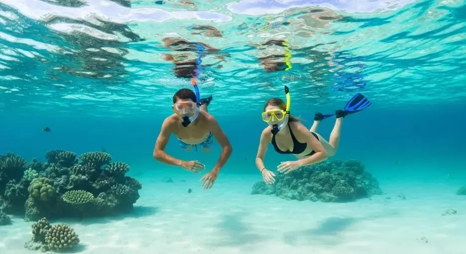 Two people snorkeling over a coral reef in clear blue tropical water, surrounded by small fish and sunlight reflecting on the surface
